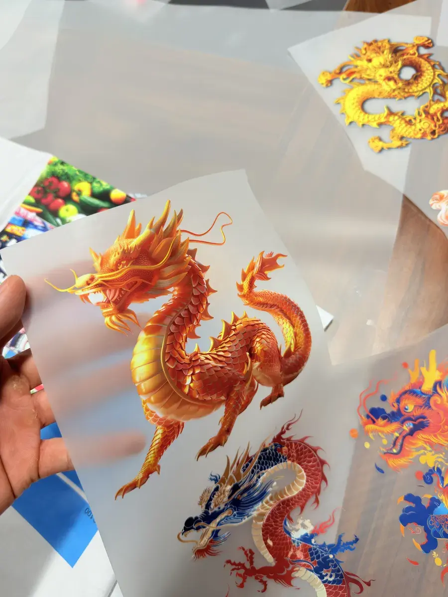Printed dragon and floral DTF transfers held up on clear PET film before pressing