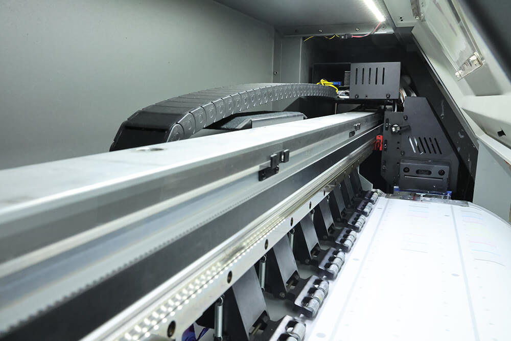 Interior de una impresora de formato ancho con viga de aluminio extruido, cadena portacables y una fila de rodillos de presión que mantiene plano el material sobre la platina