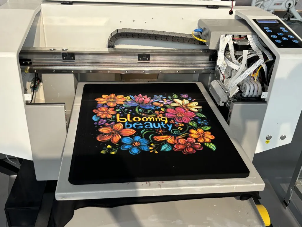 Impressora DTG aberta com uma camiseta preta na mesa e uma estampa floral multicolorida