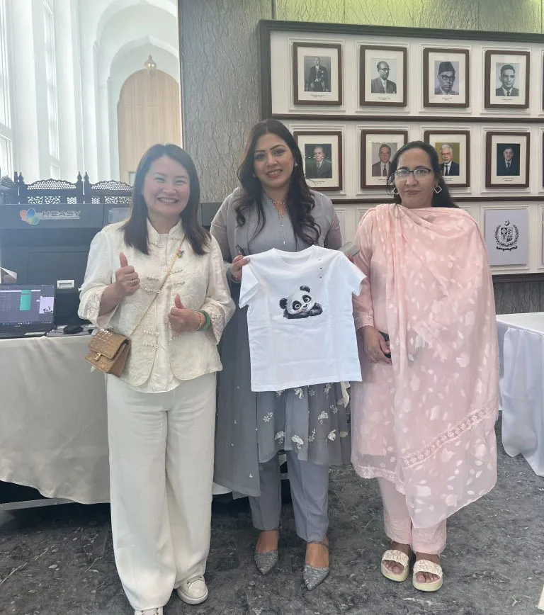 Convidados seguram uma camiseta personalizada com um panda ao lado da impressora DTG HY-4050 na Embaixada do Paquistão em Pequim